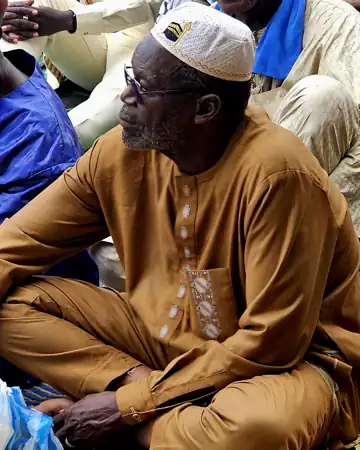 Un aîné en boubou ocre, assis en tailleur.